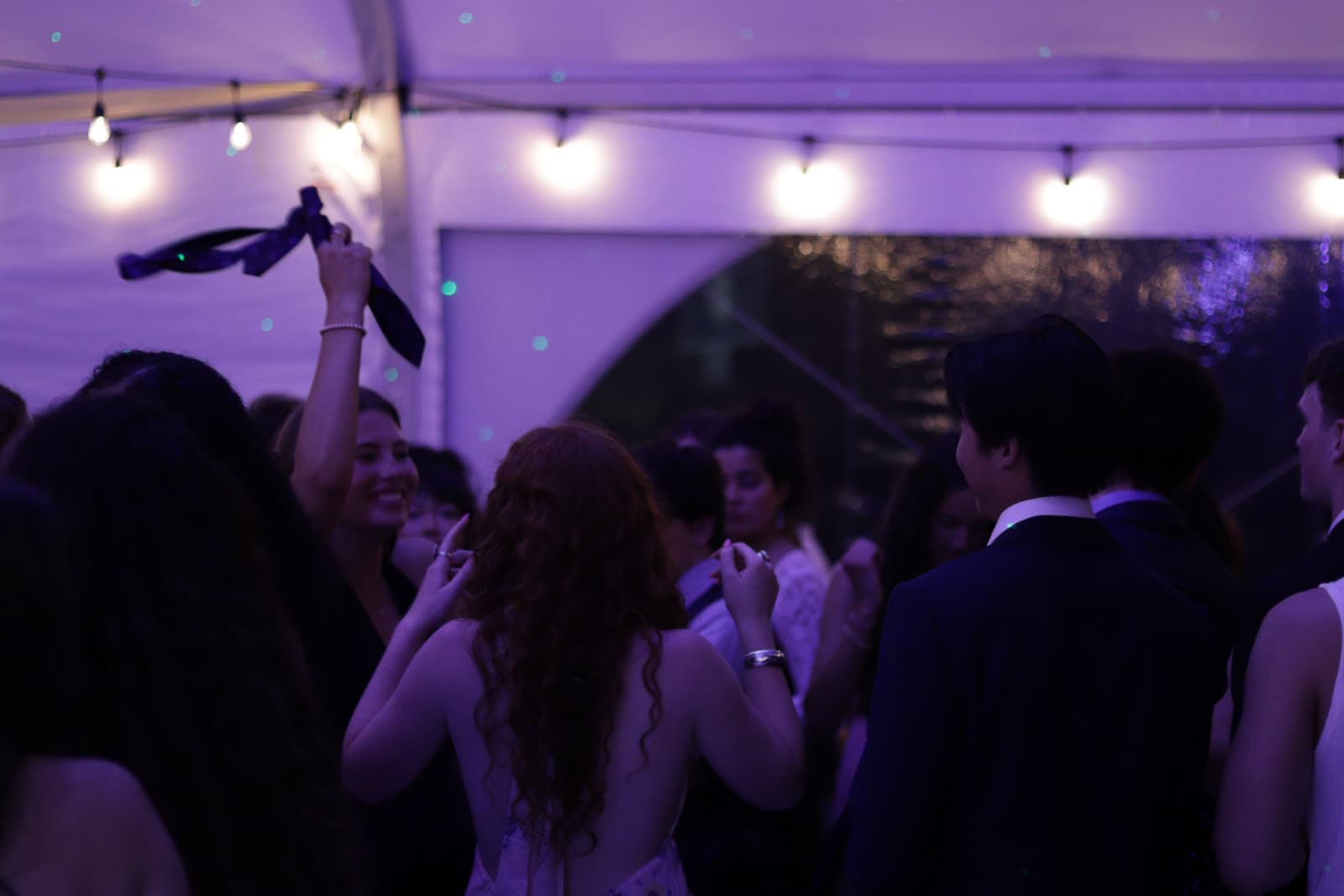Guests dancing under string lights in a tent, a tie held up in the air